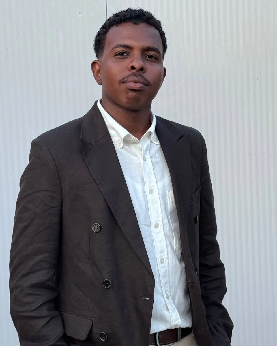 Ahmed Eltigani, in a dark linen blazer and white shirt, standing against a plain wall