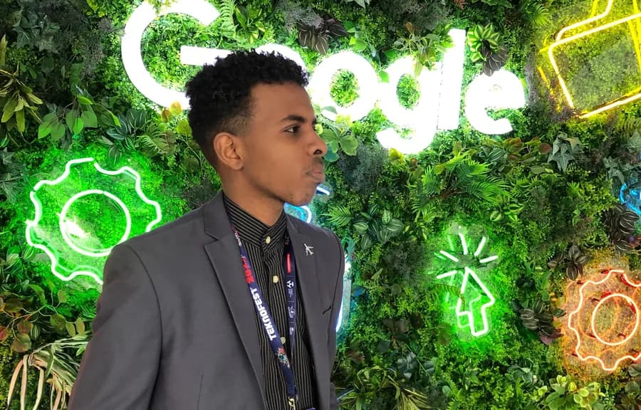 Ahmed Eltigani wearing a TEKNOFEST lanyard in front of a green wall of plants and neon icons
